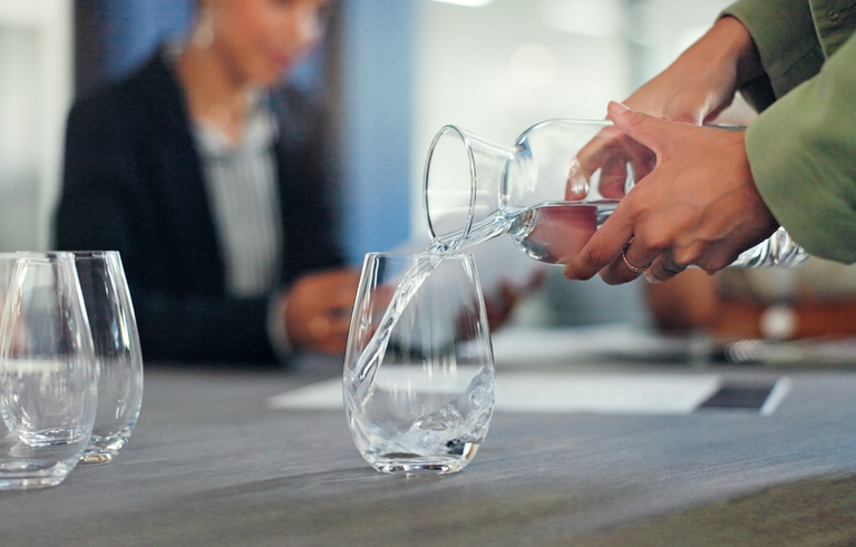 A person pours water from a glass pitcher into a glass on a table, with two empty glasses nearby and a blurred person sitting in the background.